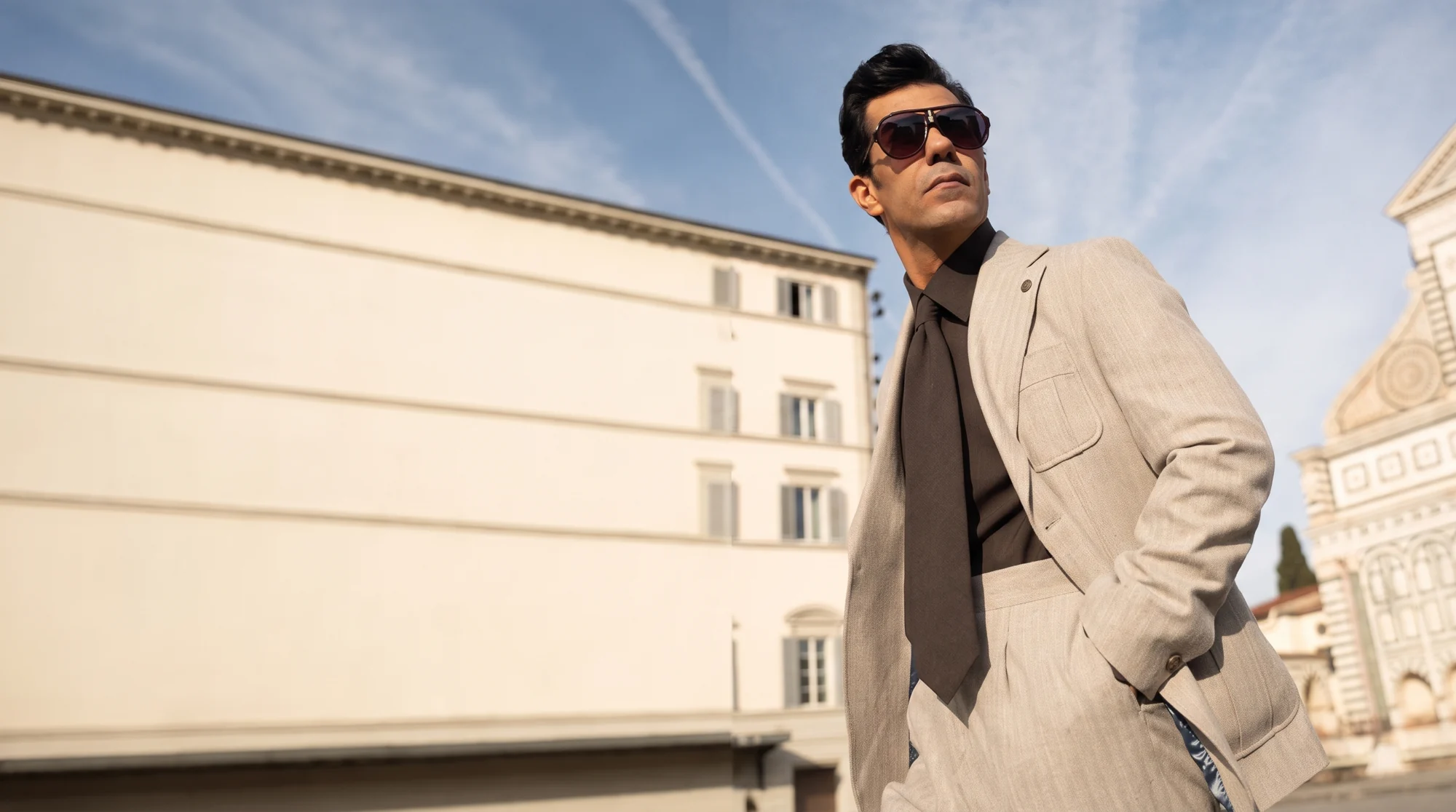 AQEEL FANTAZIA model in a beige suit, brown shirt and tie, wearing sunglasses against limestone architecture.