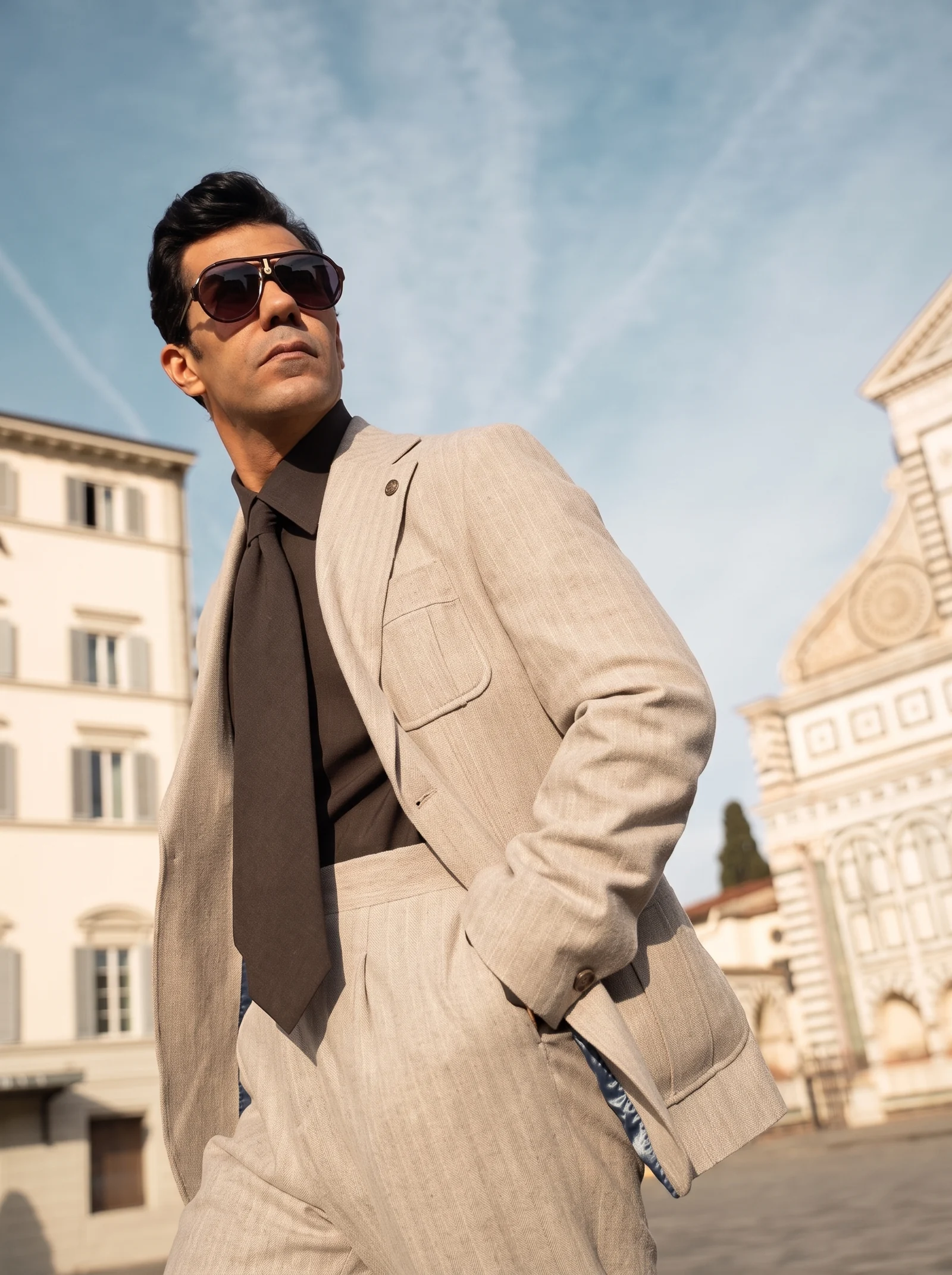 AQEEL FANTAZIA model in a beige suit, brown shirt and tie, wearing sunglasses against limestone architecture.